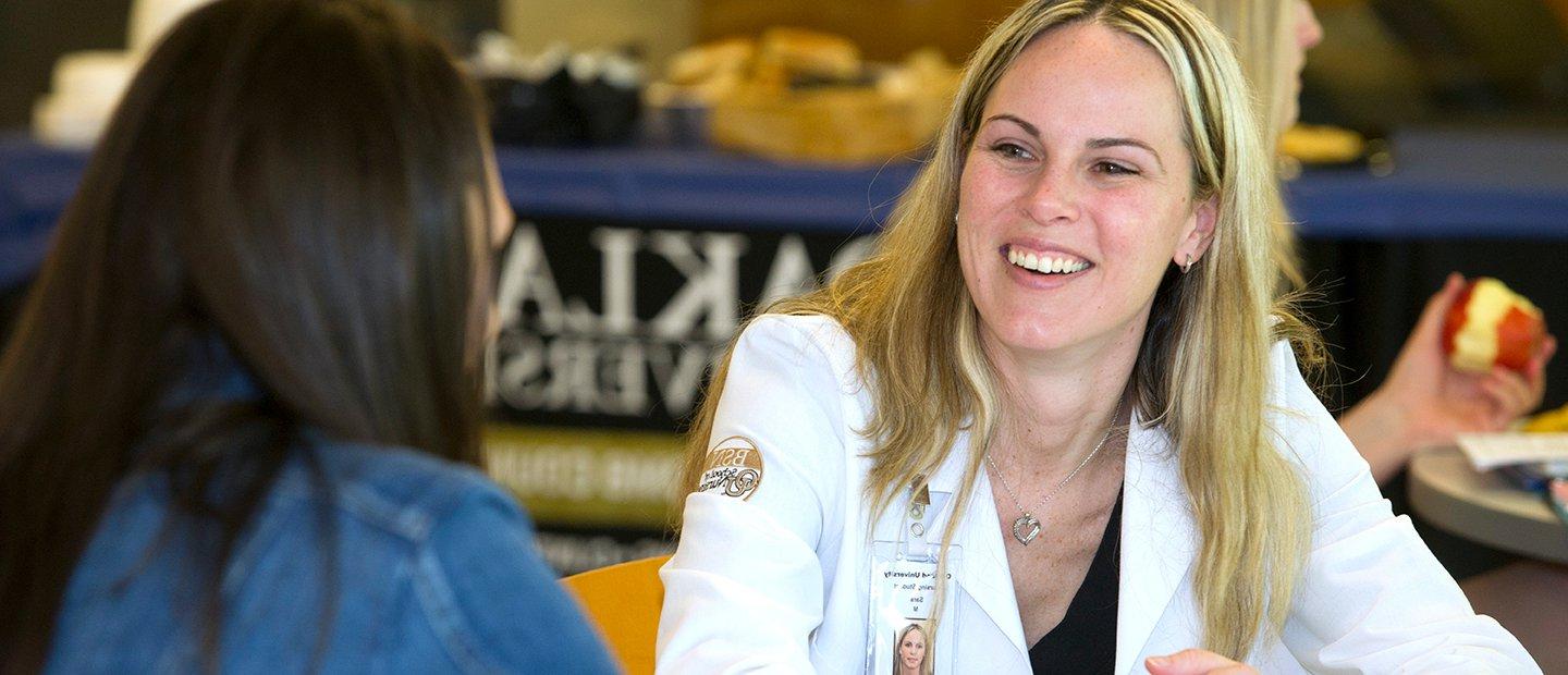 woman in a white lab coat smiling, speaking to another woman in front of her